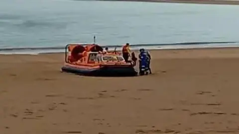 An orange RNLI hovercraft beached on a north Norfolk sandy beach. The grainy footage shows lifeboat crew helping someone on to the beach while a cluster of HM Coastguard staff in blue and white stand next to the boat.