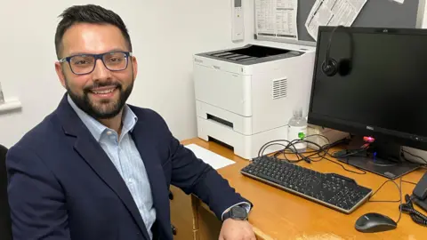 Dr Amit Sharma sitting at a desk is smiling at the camera, wearing a light blue shirt and navy jacket. He has dark hair and beard and is wearing blue framed glasses on his face.