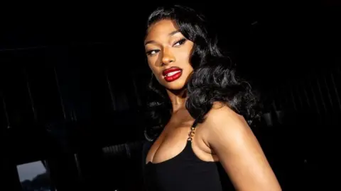 Megan Thee Stallion at a gala event in New York on 16 April 2024. Megan is a 29-year-old black woman with long wavy black hair. She wears a low-cut black dress and a bold red lipstick. She's pictured from the side, looking just past the camera over her left shoulder. The room behind her the room is dimly lit and there are champagne glasses laid out on a table.