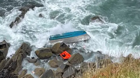 Overturned blue hull of a small boat in choppy sea next to rocks next to the shore.