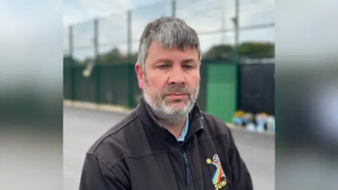 George Carden/BBC James Wilson imaginó en el campo de fútbol de Chichester City FC. James tiene cabello negro/gris y lleva una larga chaqueta negra y una camisa azul. Las alcantarillas y las cartas de las flores se pueden ver en el fondo, a pesar de estar parcialmente borrosa.