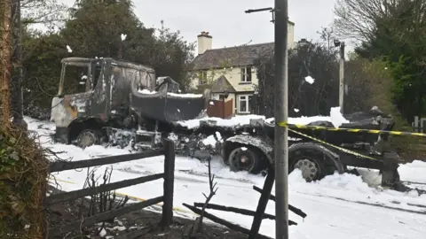 Wellington Fire Station The burned-out remains of a heavy goods vehicle can be seen in the centre of a road which is covered in white foam. There is black and yellow tape stretched between a post and the rear of the vehicle. A house can be seen in the distance behind the vehicle.