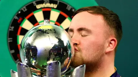 Reuters Luke Littler kisses his world champion trophy in January. He has short dark hair and stubble, a dart board can be seen behind him.