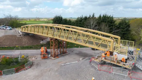 A pedestrian bridge is being installed over a road, with construction workers and equipment below.
