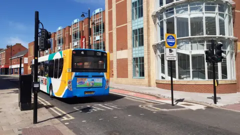 A single-decker bus driving down a street. 'Bus Gate' is written on the road and a sign with 'traffic enforcement camera' alongside a picture of a bike, bus and the word 'taxi'. Black cameras are pointing down the street.
