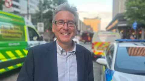 Simon Foster has grey hair and is wearing a navy suit jacket and a white shirt with blue stripes. In the background is a white, blue and yellow police car on the right, and behind that a white, red and yellow police van