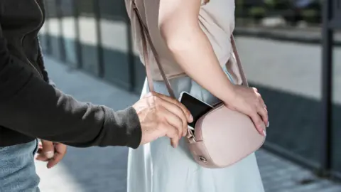 A creative image of a thief stealing a phone from a woman's handbag. The identities of both are obscured