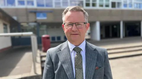 A man with grey hair, glasses and a grey suit. He stands in front of a school.