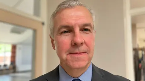 Shariqua Ahmed/ BBC Jonathan Jelley, a man with grey hair, wearing a grey suit, smiling for the camera. 