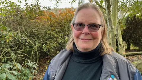 A woman with short light blonde hair wearing glasses, a black polo neck and a light grey waterproof jacket, smiling at the camera with trees and shrubbery behind her.