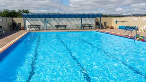 Lengthways view of a swimming pool full of water, but with no swimmers. There are benches on a partially-covered paved area at one end, and to the left a rolled up blue tarpaulin cover and some swimming aids.