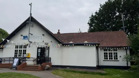 The photo shows a white building. It has a tiled roof and two Georgian style windows. There is multi-coloured bunting also hanging from it. 
