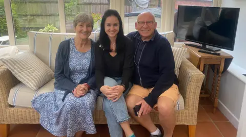 Allison, with short, grey hair, is on the left and wearing a blue floral dress and cardigan. Soffiia, with long-ish dark hair, is in the centre and has a black top and jeans on and David, with limited hair and wearing glasses, has a blue top and orange shorts. They are seated together in a conservatory on a whicker sofa.
