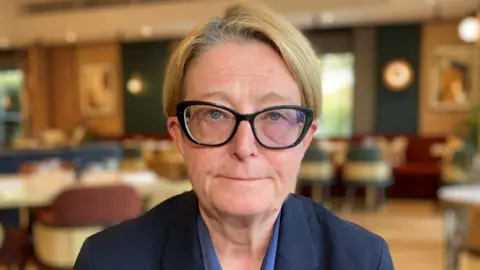 Emma Jones faces the camera wearing a serious expression. She has short, fair hair and wears large, black glasses and a blue jacket.