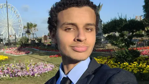 Contributed A selfie style picture of Mr Alkhatip who has short curly black hair and is wearing a dark suit with a blue shirt. He is standing in the cliff top gardens in Clacton with a ferris wheel in the background