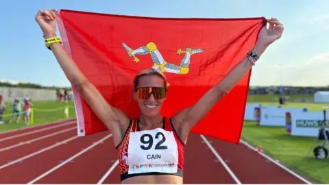 Christa Cain holds up a red Manx flag with a white grey and yellow triskelion. She wears orange sunglasses, a red crop top and a white sign on her chest, she stands on an orange running track.
