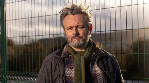 Michael Sheen looks at the camera. He is wearing a green scarf and a plaid red shirt and a coat over the top. Behind him is a waste site which is fenced off. 