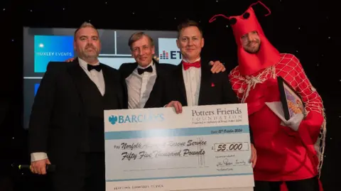 Three men in black tuxedo suits and white shirts. All have bow-ties. A man in a red lobster costume stands with them. A large cheque is being held by one of the men.