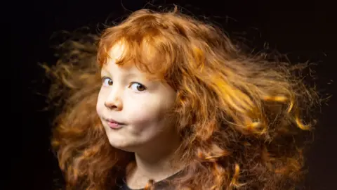 Close-up portrait of a young child with long, curly red hair flowing outward as if caught by a breeze. The child is turned slightly to one side and looking toward the camera, wearing a dark top against a dark, softly blurred background. Warm lighting highlights the texture and movement of the hair.