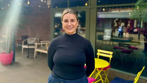 Katie Cross, a woman in a black top and blue trousers stood in front of a shop.