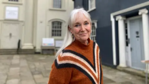 Bridport Arts Centre Ines Cavill is smiling at the camera, has blue eyes, white hair and is wearing a burnt orange jumper.