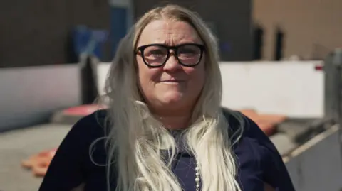 A close-up outdoor portrait of a woman facing the camera. She has long, light-coloured hair and is wearing dark-framed glasses, a dark top, and a long beaded necklace.