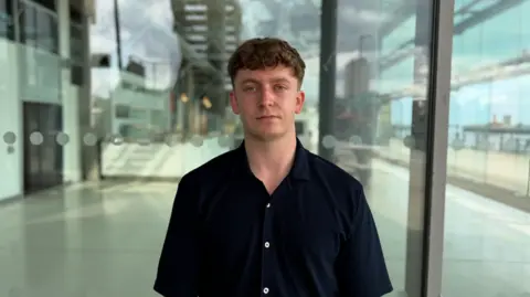 BBC/Jim Scott Labour's councillor Brandon Feeley, who is wearing a blue open-buttoned shirt, stands in front of the now closed National Glass Centre. In the background is the counter which formed part of the restaurant, and the empty restaurant space.