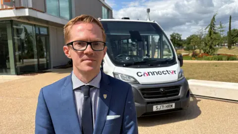 Ben Schofield/BBC Man wearing a suit and glasses stands in front of a driverless bus