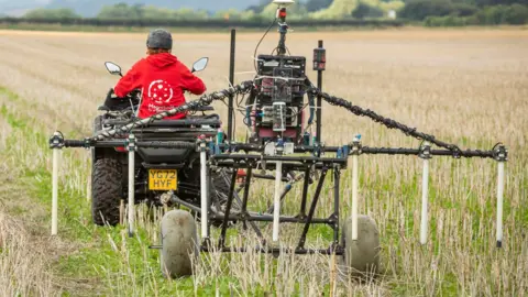 A person in a red top on a quad bike towing survey equipment across a field. The equipment is made up of several metal poles with some dangling over the field and mounted on two wheels.