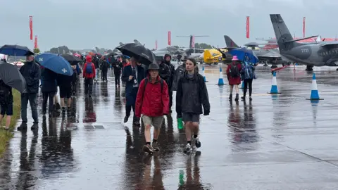 Groups of people walking on the runway at RIAT wearing coats and holding umbrellas. They look very wet. There are planes in the background and reflections of the people in the puddles.