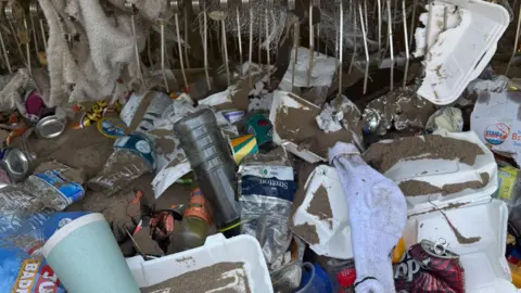 Rubbish which has been gathered in the tractor. There is takeaway cups, boxes, plastic bottles, cans and a grey sock all covered in sand. A towel and a cardboard box sits in the teeth of the comb on the tractor. 