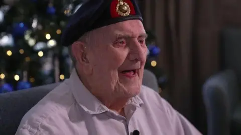 An elderly veteran wearing a navy beret with a green and gold logo at the front. He is smiling with his mouth open and wearing a long-sleeved light pink shirt. He is sat in a grey chair and the background is blurred but you can see fairy lights from a Christmas tree.