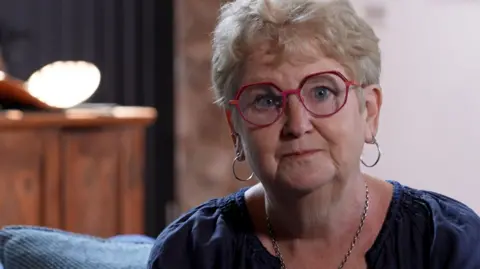 Dr Jane Dickson, a woman with short blonde hair and large red glasses, sits on a blue sofa, looking into the camera. She is wearing a dark blue top, silver hoop earrings and a silver necklace