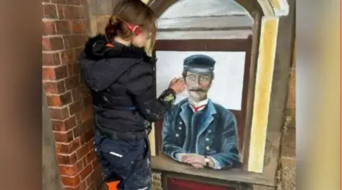 A woman wearing a black coat and black trousers, with splattering a paint, is standing over a painting with a brush in hand. The painting depicts a Victorian-era train station ticket officer sitting with his hands on a ticket office ledge.