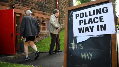 A picture of voters heading in and out of a polling station, with a polling place sign in the foreground 