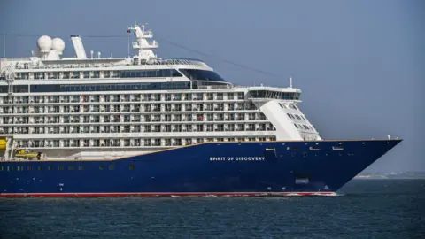 A large cruise ship with a blue hull saying 'Spirit of Discovery' on the site, in a stretch of open water. It's a sunny day and the sky is blue.