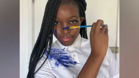 Joy Nzau A woman with long braids is wearing a white school shirt with blue paint on the left shoulder. She is holding a paintbrush to her nose.