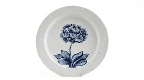 Woolley and Wallis A image of a white plate with a blue flowering auricula painted on it. It is propped up and in front of an all-white background.
