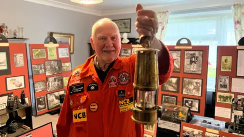 A man wearing orange mining overalls and with grey hair is holding a miner's lamp up in a room where behind him is a collection of mining pictures and artefacts.