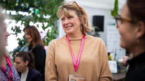 Nicola Tuxworth, in focus, smiling to someone off camera, wearing a beige jumper and a pink lanyard, with dusty blonde hair and glasses on top of her head, surrounded by people out of focus