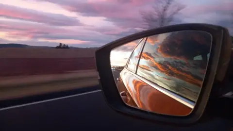 A car mirror reflecting a purple, pink and orange sunset with a landscape in the background