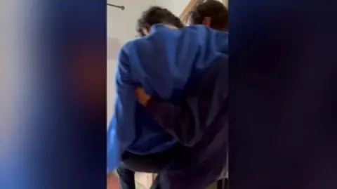 Family handout A man wearing a dark blue jumper and black trousers carries his disabled son up the stairs.