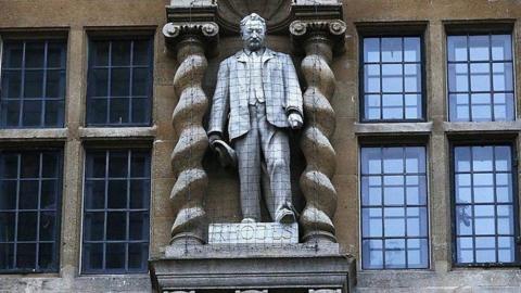 A statue of a man attached to a building, with columns each side of him. To his left and right are windows. There is black netting covering him. Beneath his feet is a plaque that says "Rhodes".