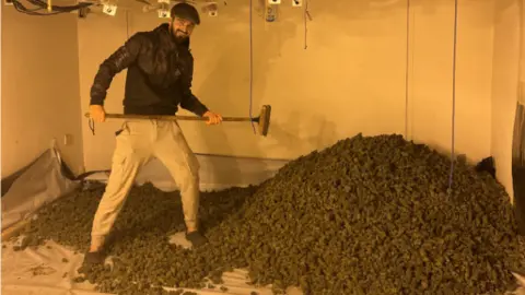 Greater Manchester Police Man stands beside huge pile of cannabis smiling and carrying a broom.