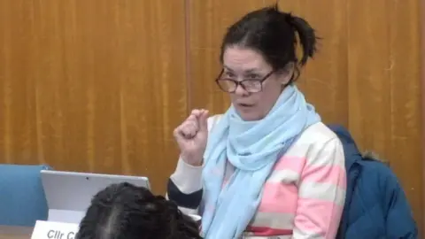 Warwickshire County Council Clare Golby, sitting in a council chamber, she is wearing a blue scarf and a jumper with large white and pink stripes