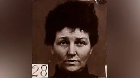 Black and white mugshot of a woman with dark hair piled on top of her head. She has a high collar around her neck and is looking slightly above the camera. There is a number - 28 - in the bottom left hand corner. 