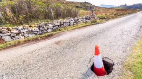 An orange traffic cone sits in a deep pothole on a country single-track road. there is a small stone wall on the far side of the road, which is bordered by a grass verge with hills in the background