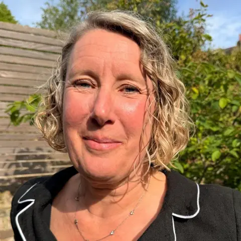 Sarah Branson has wavy light hair and is wearing a dark jacket, she is standing in a sunlit garden with a wooden fence and green foliage softly blurred in the background.