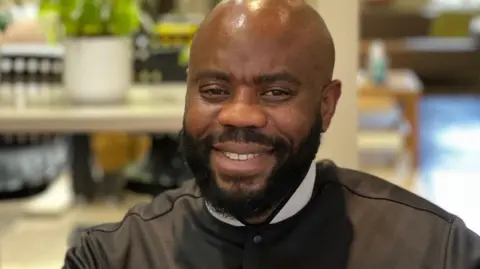 Jeff Ukiri with a dark brown beard wears a long-sleeved brown top with white collared top underneath. He is sitting in an office. He is smiling. 