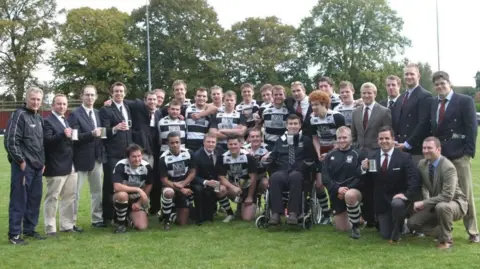 Stratford upon Avon rugby players and staff stand together on the pitch for a team photo in 2012 with Gareth Rees in the front in his wheelchair.